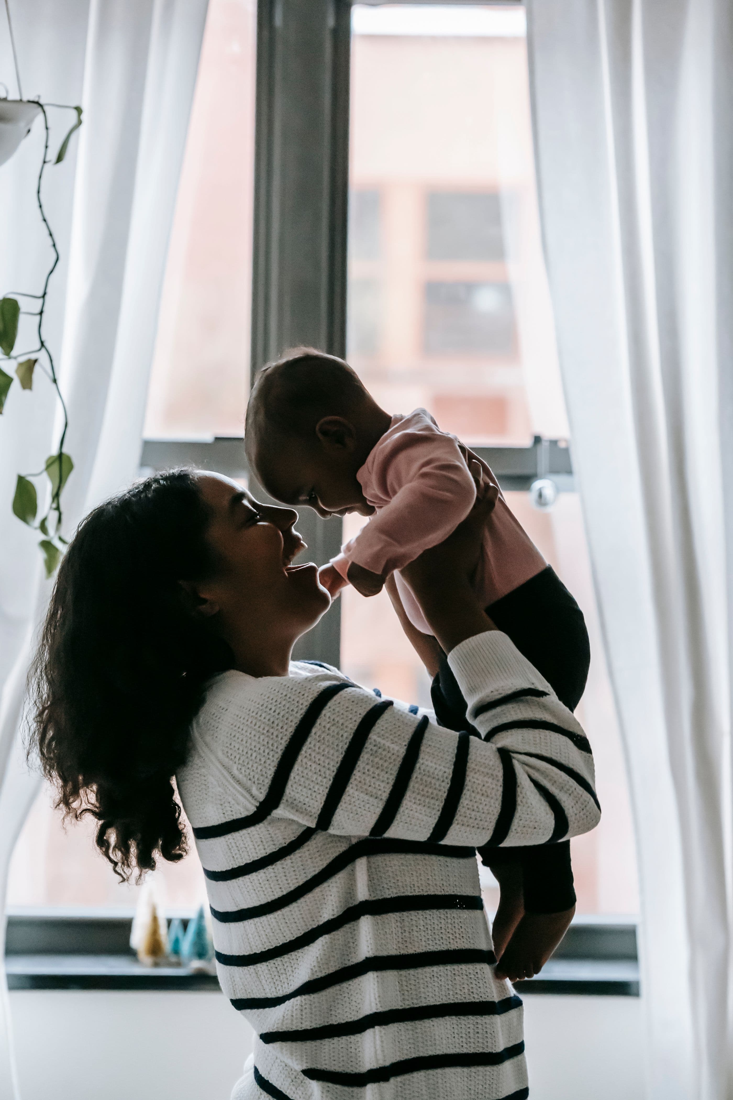 A parent lifting their baby in the window light of a city flat.